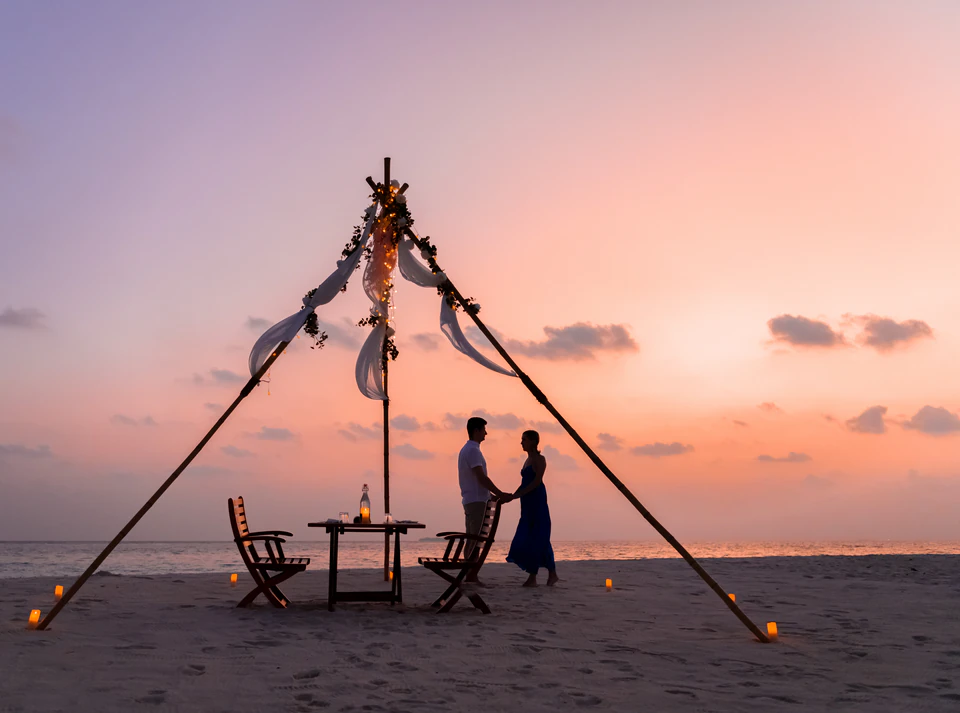 Private beach dinner setup with a couple beneath a decorated canopy at sunset
